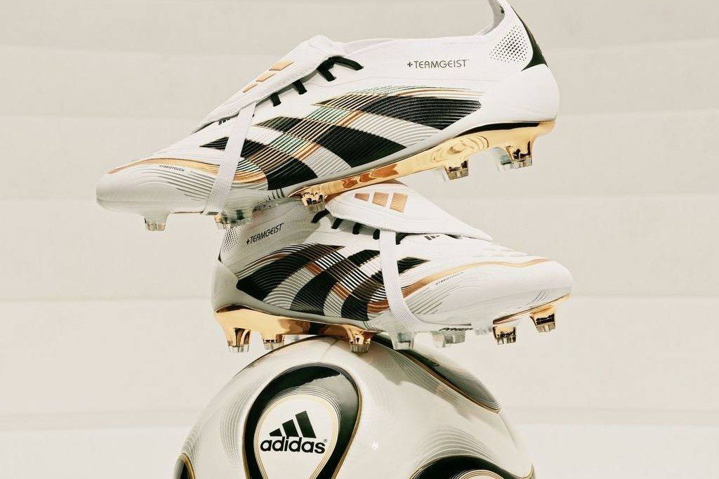 Botines Adidas blancos con franjas negras y tapones dorados sobre una pelota
